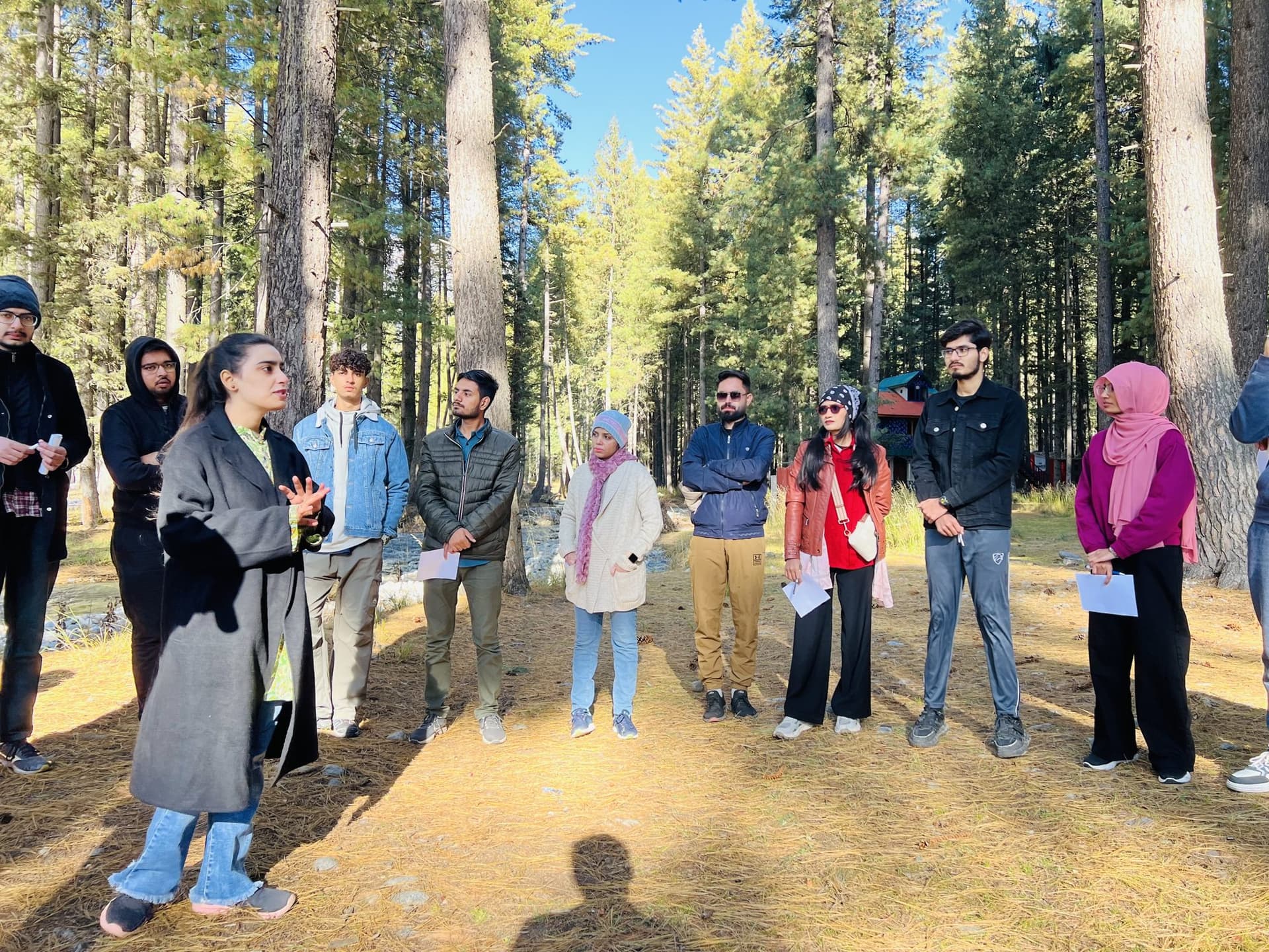 Lalain talking to a circle of young people standing in a pine forest