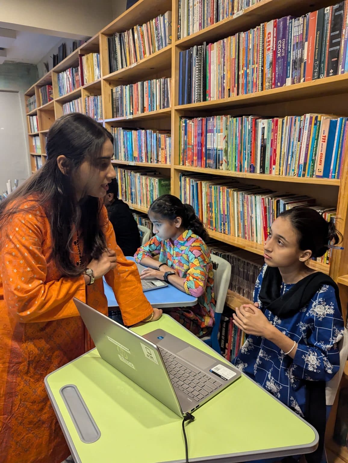 Lalain in orange, leaning in to help two students at a laptop during a Dialogue Academy course